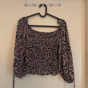 Blommig topp med långa ärmar - Säljer en superfin blommig topp funkar som vanlig och off shoulder. Den har långa ärmar och en smockad design som ger en tight passform. Perfekt för både vardag och fest!