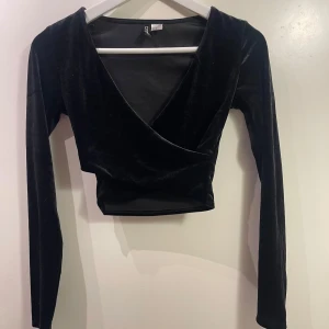 Svart omlottopp från H&M Divided - Snygg svart omlottopp i sammet från H&M Divided. Den har en elegant v-ringning och långa ärmar, perfekt för en kväll ute eller en fest. Toppen är i storlek XS. 