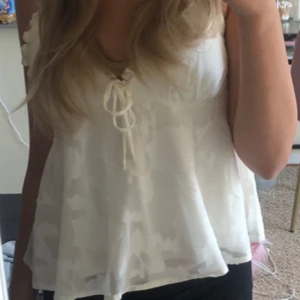 Vit blommig linne med knyt - Säljer en söt vit linne med blommigt mönster och knytning framtill. Toppen har smala axelband och en luftig passform, perfekt för sommardagar. Den är i mycket bra skick och ger en romantisk touch till din outfit.