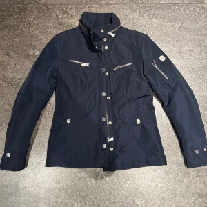 Mörkblå jacka från Snoot - Snygg mörkblå jacka från Snoot i storlek S. Jackan har en stilren design med flera dragkedjefickor och knappar. Perfekt för höst och vår med sitt quiltade foder som håller dig varm. Passar både till vardags och lite finare tillfällen.