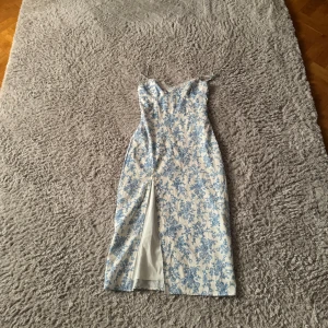 Blommig klänning med slits och spets  - Säljer en superfin vit klänning med blått blommönster. Den har smala axelband och en snygg slits framtill. Perfekt för sommarens fester eller en mysig vårdag. Klänningen är i bra skick och har en elegant passform.