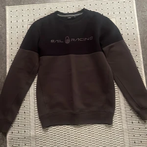 Svart och grå tröja från Sail Racing - Säljer en snygg svart och grå tröja från Sail Racing i storlek S. Tröjan har en stilren design med långärmade ärmar och en bekväm passform. Perfekt för både vardag och fritid. Den är i mycket bra skick och redo för en ny ägare!