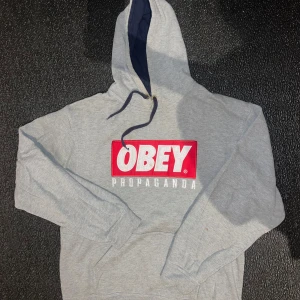 Grå hoodie från Obey - Säljer en grå hoodie från Obey med en stor röd och vit logga på framsidan. Den har en klassisk passform med långa ärmar och en justerbar huva. Perfekt för en avslappnad stil eller streetwear-look. Hör av dig vid intresse!