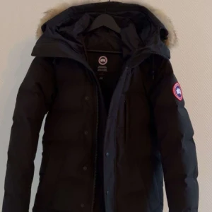 Svart dunjacka från Canada Goose - Säljer en svart dunjacka från Canada Goose med pälsdetaljer på huvan. Perfekt för kalla vinterdagar. Jackan har en dragkedja och är i mycket bra skick. Den är långärmad och har det klassiska Canada Goose-märket på ärmen.