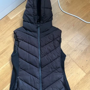 Svart dunväst från Zara - Säljer en svart dunväst från Zara i mycket bra skick. Västen har en dragkedja framtill och en praktisk huva. Perfekt för höst och vår när du vill hålla dig varm utan att bära en jacka. Passar bra till både vardag och lite mer avslappnade tillfällen.