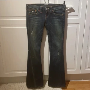 Mörkblå bootcut jeans - Säljer ett par snygga mörkblå bootcut jeans från True Religion. De har en låg midja och coola slitningar på framsidan. Bakfickorna har snygga broderade detaljer och knappar. Perfekta för en avslappnad men stilren look!