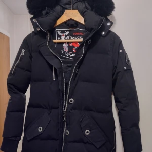 Svart dunjacka från Moose Knuckles - Säljer en snygg svart dunjacka från Moose Knuckles i  bra skick. Observera att storleken är XS/TP (tre petite)  och att den passar smal tjej runt 160 cm eller uppskattningsvis ungdom 158/164. Jackan köptes på Plick för två veckor sedan och var felmärkt i annons som S och unisex. Tror att jackan kan vara äkta men tidigare ägare har tappat tagg och lagat sömmen, se bild 5. Jackan har en pufferstil med dragkedja och knappar, samt en mysig huva med pälsdetaljer. Perfekt för vintern! 
