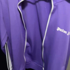 Lila jacka från Palm Angels - Säljer en snygg lila jacka från Palm Angels. Den har en dragkedja framtill och vita detaljer längs ärmarna. Perfekt för en cool och avslappnad stil. Passar både till vardags och fest!