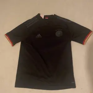 Säljer en snygg svart Tyskland fotbollströja från Adidas. Den har korta ärmar med röd och gul detalj vid ärmsluten. Perfekt för fotbollsfans som vill ha en stilren look på planen eller till vardags. Tröjan är i bra skick och redo för nya äventyr!