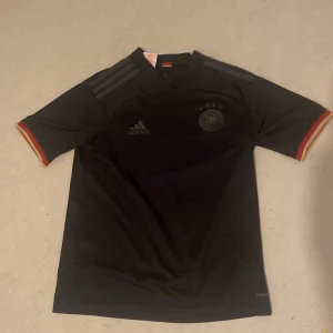 Svart Tyskland fotbollströja från Adidas - Säljer en snygg svart Tyskland fotbollströja från Adidas. Den har korta ärmar med röd och gul detalj vid ärmsluten. Perfekt för fotbollsfans som vill ha en stilren look på planen eller till vardags. Tröjan är i bra skick och redo för nya äventyr!