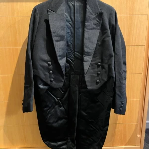 Svart frack med byxor - Säljer en elegant svart frack med matchande byxor. Fracken har klassiska slag och knappar framtill, perfekt för formella tillfällen. Byxorna är i samma stilrena svarta färg. Passar utmärkt för bal eller fest. Skick: mycket bra skick.