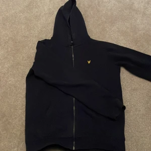 Mörkblå hoodie från Lyle & Scott - Säljer en snygg mörkblå hoodie från Lyle & Scott i storlek 14/15 år. Den har en dragkedja framtill och en liten gul logga på bröstet. Perfekt för höst och vår, och den är supermjuk och bekväm. Passar både till skolan och fritiden!