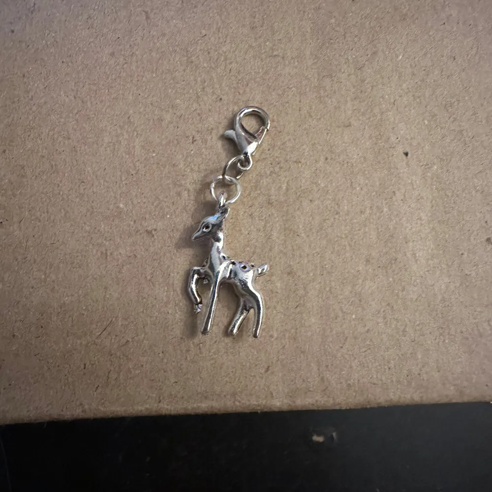 Söt liten berlock i form av ett rådjur, perfekt för att fästa på ett armband eller halsband. Den är silverfärgad och har en praktisk karbinhake för enkel fastsättning. Perfekt för djurälskare eller som en fin detalj på ditt smycke.. Asusteet.