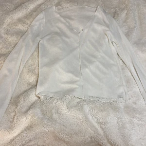 Vit långärmad topp - Säljer en stilren vit långärmad topp med v-ringning. Toppen har en enkel design med vågig kant vid nederdelen som ger en fin detalj. Den är genomskinlig, och passar bra till lite mer off shoulder. 