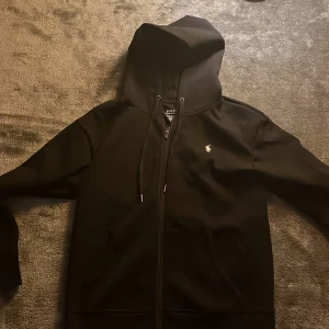 Svart hoodie från Ralph Lauren - Snygg svart hoodie från Ralph Lauren med dragkedja och justerbar huva. Den har en liten broderad logga på bröstet och är i en slim passform. Perfekt för en stilren och bekväm look.