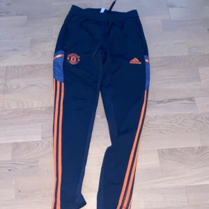 Svarta träningsbyxor från Adidas med orange detaljer - Snygga svarta träningsbyxor från Adidas med orange ränder längs sidorna och Manchester United-logga. Perfekta för fotbollsträning eller annan sportaktivitet. Byxorna har en bekväm passform och elastisk midja.
