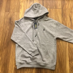 Grå hoodie från Polo Ralph Lauren - Säljer en stilren grå hoodie från Polo Ralph Lauren med det klassiska logotypbroderiet på bröstet. Tröjan är helt ny sitter som en M men står L i tröjan.