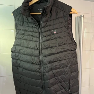 Svart dunväst från Gant - Snygg svart dunväst från Gant med dragkedja framtill. Perfekt för kyligare dagar och enkel att matcha med andra plagg. Storlek 170/S men passar mig som är 180 cm och väger 75 kg. Använd väldigt lite och är i mycket bra skick.
