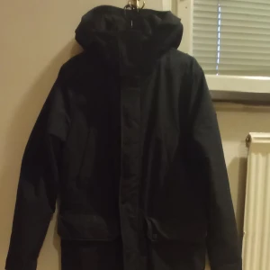 Mörkblå jacka från J.Lindeberg - Snygg jacka från J.Lindeberg med dragkedja och knappar. Perfekt för kyligare dagar med sin långa design och praktiska fickor. Jackan har långa ärmar och en stilren look. Nypris 7 800kr