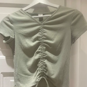 Grön ribbad topp från Lindex - Snygg grön ribbad topp från Lindex med v-ringning och knytning framtill. Perfekt för en avslappnad stil. Kortärmad design som passar bra till både jeans och kjol.