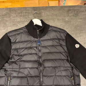 Svart dunväst med ärmar - Snygg svart moncler cardigan med dragkedja och stickade ärmar. Perfekt för kyligare dagar med sin  stilrena look. Både qr kåd och nfc-scan fungerar. Bara höra av dig vid frågor. Kan gå ner i pris vid smidig affär 