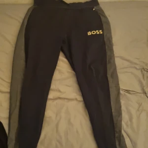 Boss byxor - Snygga Blå mjukisbyxor från Boss med grå sidopaneler. Perfekta för en avslappnad stil. Byxorna har en elastisk midja och en logotyp på ena benet.