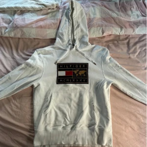 Vit hoodie från Tommy Hilfiger - Säljer en stilren vit hoodie från Tommy Hilfiger med en stor logotyp på bröstet. Hoodien har en klassisk känguruficka och justerbar huva med dragsko. Perfekt för en avslappnad look.