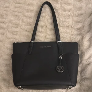 Svart axelväska från Michael Kors - Elegant svart axelväska från Michael Kors i skinn med silverdetaljer. Väskan har dubbla handtag och en dekorativ MK-berlock. Perfekt för att bära dina dagliga nödvändigheter med stil.