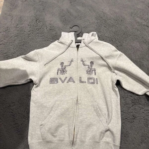 Bvaldi hoodie - Säljer en grå hoodie med dragkedja och tryck på framsidan. Perfekt för en avslappnad stil. Den har långa ärmar och en bekväm passform.