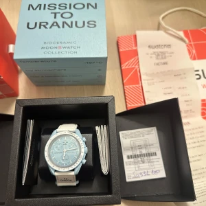 NY klocka från Swatch x Omega mission to Uranus  - Säljer en stilren blå klocka från Swatch x Omega's Bioceramic Moonswatch Collection, 'Mission to Uranus'. Klockan har en ljusblå urtavla och vit boett med matchande armband. Perfekt för den som gillar unika och moderna accessoarer. Helt ny med allt og och swatch påsen. Kvitto finns från butiken.