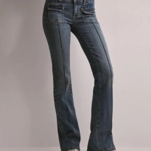 Blå bootcut jeans från Nelly - Snygga blå bootcut jeans från Nelly med låg midja och dubbla knappar. Perfekta för en avslappnad stil med sina framträdande fickor och klassiska jeanslook.