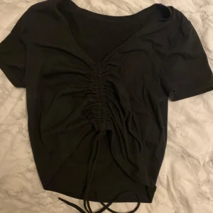Svart kortärmad topp med snörning - Snygg svart topp med korta ärmar och v-ringning. Den har en snörning framtill som ger en fin detalj. Perfekt för en stilren look.