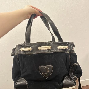 Svart handväska från Juicy Couture - Snygg svart handväska från Juicy Couture med ett hjärta och bokstaven 'J' på framsidan. Väskan har vita detaljer och ett mönstrat innerfoder med Juicy Couture-loggan. Perfekt för att bära dina dagliga nödvändigheter med stil.