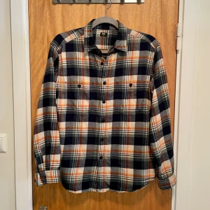 Rutig skjorta från Uniqlo - Snygg rutig flannelskjorta från Uniqlo i blått, orange och vitt. Skjortan har långa ärmar och knappar framtill. Perfekt för en avslappnad stil. Använd bara ett fåtal gånger. Axelvidden är 46 cm och längden är 69 cm. Paketeras och skickas ca 1-3 dagar efter köp. Frågor? Skicka ett dm!