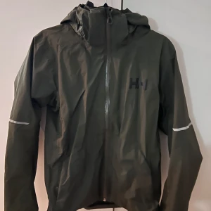 Grön vindjacka från Helly Hansen - Snygg och funktionell grön vindjacka från Helly Hansen med huva och dragkedja. Perfekt för blåsiga dagar och utomhusaktiviteter. Jackan har långa ärmar och reflekterande detaljer på ärmarna.