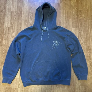 Blå hoodie från Urban Outfitters - Säljer en snygg blå baggy hoodie från Urban Outfitters med ett coolt månmotiv och stjärnor på bröstet och ryggen. Tröjan har en stor ficka fram och justerbar huva. 