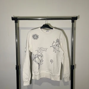 Vit tröja från Zara - Säljer en vit tröja från Zara med unika tryck av sol och ansikten. Tröjan har långa ärmar och en rund halsringning. Perfekt för en stilren och konstnärlig look.