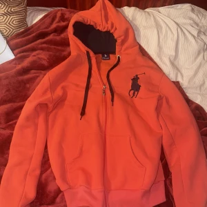 Orange huvtröja från Polo Ralph Lauren - Snygg orange huvtröja från Polo Ralph Lauren med dragkedja och svart broderad logga på bröstet. Tröjan har svarta snören i huvan och är perfekt för en avslappnad stil.