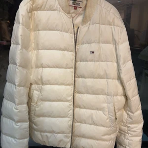 Vit pufferjacka från Tommy Hilfiger - Säljer en stilren vit pufferjacka från Tommy Hilfiger. Jackan har en dragkedja framtill och det klassiska logotypmärket på bröstet. Perfekt för kyligare dagar med sin quiltade design.