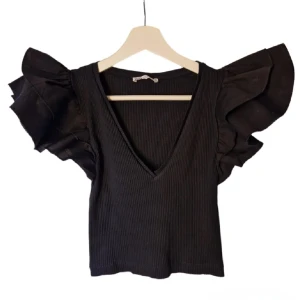 Svart ribbad topp från Zara - Snygg svart ribbad topp från Zara med volangdetaljer på ärmarna och en djup v-ringning. Perfekt för att ge en stilfull touch till din outfit.