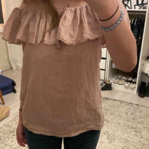 Randig offshoulder topp med volang - Säljer en söt randig offshoulder topp i rosa/beige och vit med volangdetaljer. Perfekt för sommardagar 