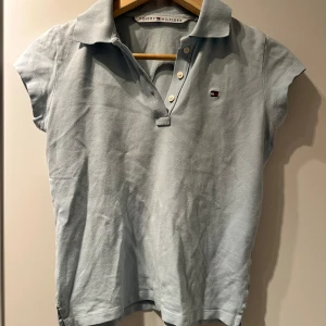 Ljusblå pikétröja från Tommy Hilfiger - Snygg ljusblå pikétröja från Tommy Hilfiger med klassisk krage och knappar framtill. Tröjan har korta ärmar och en liten logga på bröstet. Perfekt för en avslappnad stil eller sthlm stil. Dock har den ett hål på vars en sida där nere som är enkla och fixa med nål och tråd.