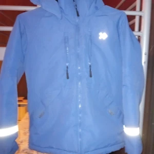 Blå jacka från Isbjörn of Sweden - Säljer en blå jacka från Isbjörn of Sweden med reflekterande detaljer och dragkedja. Jackan har en huva och är perfekt för kyligare väder. Den är designad för att hålla dig varm och synlig i mörkret.