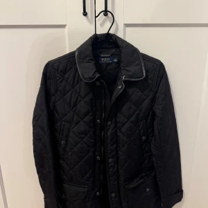 Svart quiltad jacka från Polo Ralph Lauren - Snygg svart quiltad jacka från Polo Ralph Lauren med knappar och dragkedja. Jackan har en klassisk krage och två fickor framtill. Perfekt för kyligare dagar.