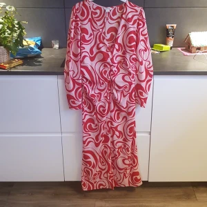 Röd och vit mönstrad klänning från Monki - Säljer en färgglad klänning från Monki med ett unikt rött och vitt mönster. Klänningen har långa ärmar och en v-ringad design med knytning framtill. Perfekt för att sticka ut med stil!