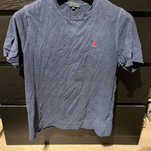 Blå t-shirt från Polo Ralph Lauren - Snygg blå t-shirt från Polo Ralph Lauren med det klassiska röda broderade logotypen på bröstet. Perfekt för en avslappnad stil. Gjord i mjuk bomull och har korta ärmar. Inköpt från Kidsbrandstore. Storlek är barnstorlek L, 14-16 år och motsvarar ungefär storlek S i herrstorlek. 