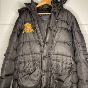 Svart dunjacka från Polo Ralph Lauren - Snygg svart dunjacka från Polo Ralph Lauren med broderad logga på bröstet. Jackan har en praktisk dragkedja och knappar framtill samt flera fickor. Perfekt för kalla dagar med sin quiltade design och långa ärmar.
