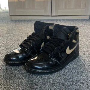 Air Jordan 1 High Black Metallic Gold - Svart-guld-glansiga Jordan 1s i storlek 44,5. Små repor finns här och där över paret och en större defekt från på högra skon (se bild 4). Köpta på Sneakershyllan i Stockholm. Box eller originalsnören finns ej. Pris kan diskuteras och hör gärna av dig om du har några frågor.