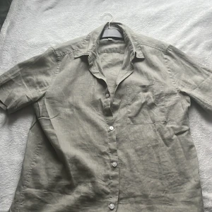Grön kortärmad linneskjorta - Säljer en stilren beige kortärmad skjorta i linne. Skjortan har knappar framtill och en klassisk krage. Perfekt för varma dagar med sin lätta och luftiga känsla.