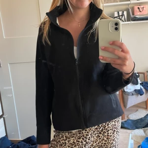 Svart fleecejacka med dragkedja - Säljer en svart fleecejacka med dragkedja framtill. Jackan har en enkel design med långa ärmar och en hög krage. Perfekt för kyligare dagar.